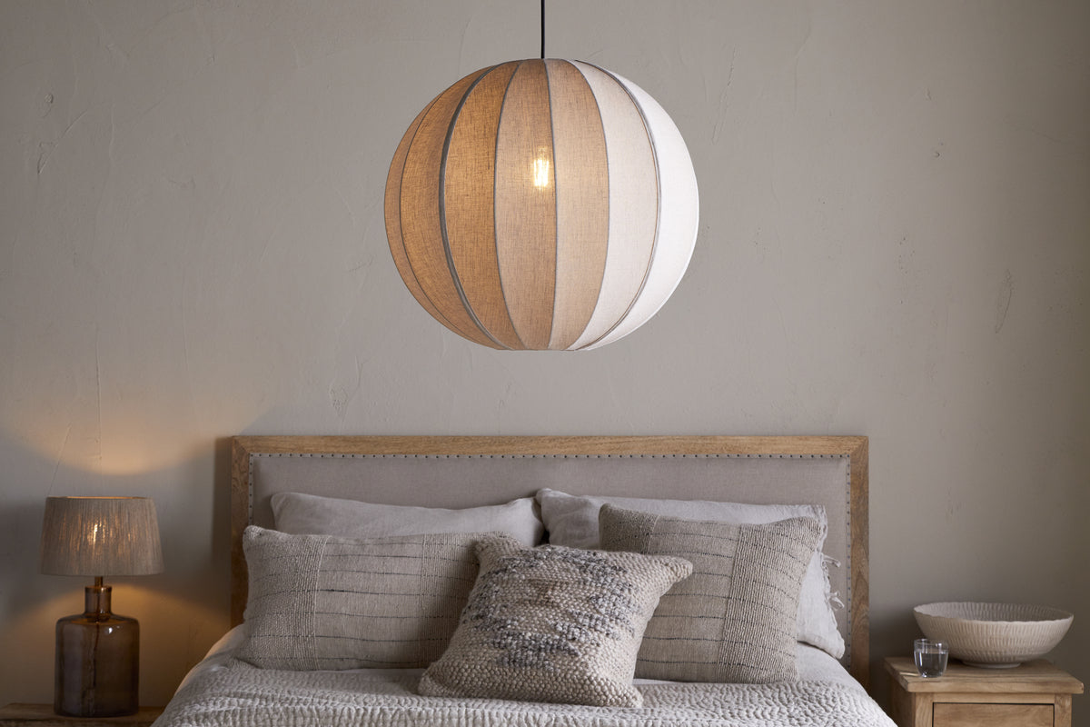 A neutral-toned bedroom showcases a bed with pale bedding, textured pillows, a wooden headboard, a small wooden nightstand with a ceramic bowl, and the nkuku Dharini Linen Lampshade - Natural hanging above.