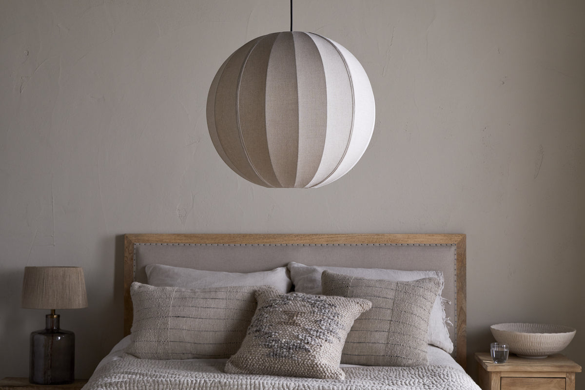 A cozy bedroom with a wooden headboard, light gray bedding, and textured pillows. Above the bed hangs the nkuku Dharini Linen Lampshade - Natural. A wooden nightstand with a small lamp and decorative bowl sits against the light gray wall.