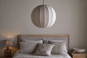 A neatly made bed with neutral bedding and textured pillows sits against a pale wall. Above, the nkuku Dharini Linen Lampshade in Natural hangs as the bedroom light. A wooden nightstand on the right holds a bowl; the left one has a lamp.
