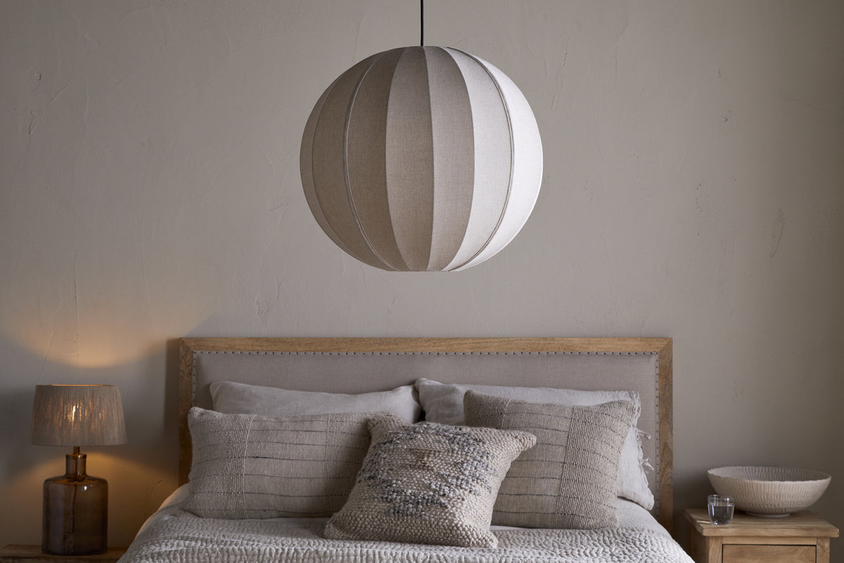 A neatly made bed with neutral bedding and textured pillows sits against a pale wall. Above, the nkuku Dharini Linen Lampshade in Natural hangs as the bedroom light. A wooden nightstand on the right holds a bowl; the left one has a lamp.