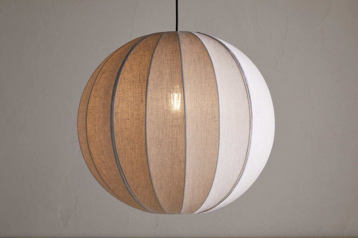 The Dharini Linen Lampshade - Natural by nkuku features a spherical linen shade with beige and white panels, suspended from a black cord against a light gray wall, softly illuminating your bedroom with its warm, textured glow.