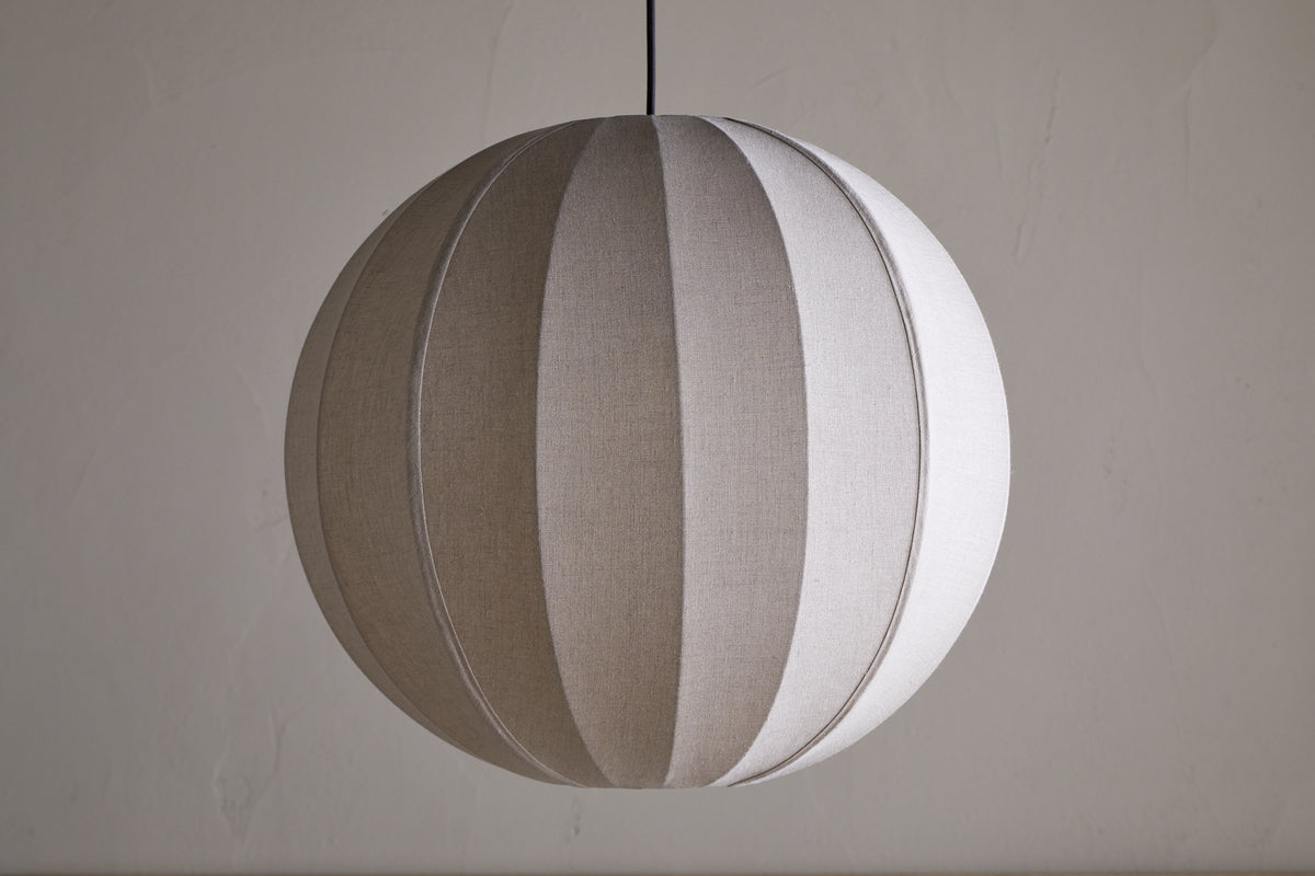 The nkuku Dharini Linen Lampshade - Natural features a round silhouette with vertical, alternating light and dark linen panels, suspended from a black cord. Its woven texture suits any elegant setting, ideal for bedroom ambiance.