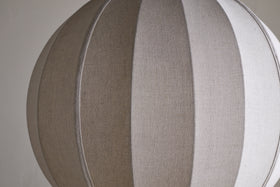 The Dharini Linen Lampshade - Natural by nkuku features a spherical, beige, textured linen design with vertical seams for a segmented look. Its simple backdrop highlights the lampshade’s elegant structure—perfect for bedroom lighting.