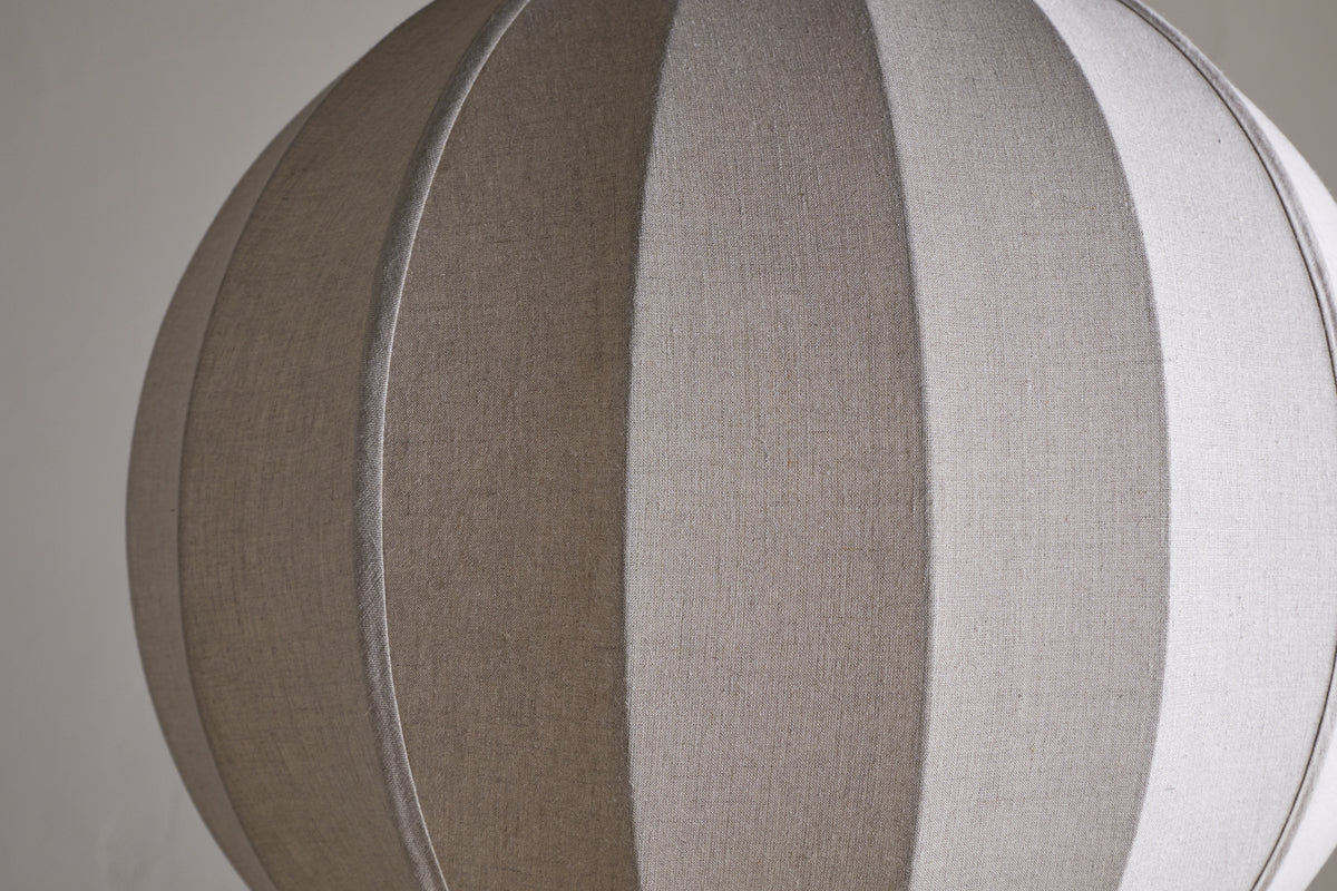 The Dharini Linen Lampshade - Natural by nkuku features a spherical, beige, textured linen design with vertical seams for a segmented look. Its simple backdrop highlights the lampshade’s elegant structure—perfect for bedroom lighting.