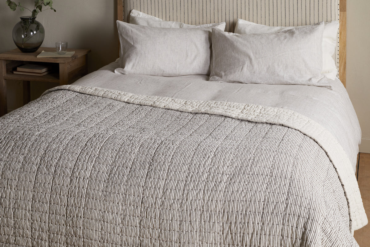 A neatly made bed with light beige and white linens, two pillows, and the nkuku Dipti Cotton & Linen Block Print Bed Quilt in charcoal. A wooden headboard and a small nightstand with a glass vase and water sit beside the bed.