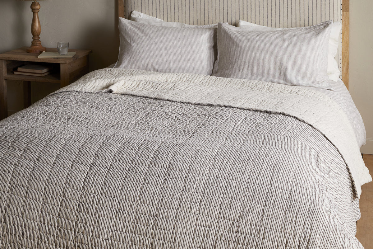 The nkuku Dipti Cotton & Linen Block Print Bed Quilt in Charcoal adds comfort and style, featuring a soft cotton linen blend with a subtle charcoal stripe design—perfect for cozying up in bed.