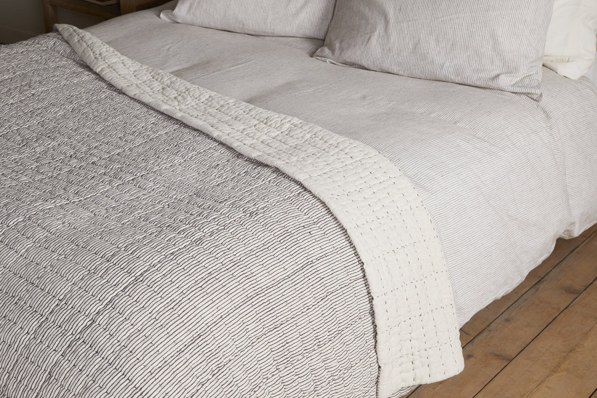 A neatly made bed features the nkuku Dipti Cotton & Linen Block Print Bed Quilt in Charcoal. Its subtle block print and cozy cotton-linen blend bring a minimalist, textured look that complements the room’s wooden floor.
