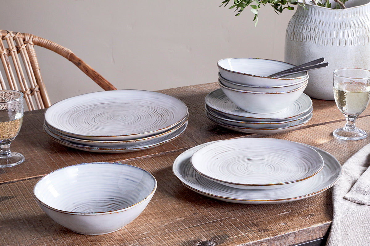 A rustic wooden table set with the nkuku Dhanuk Ceramic Dinnerware Set (Set of 12), featuring subtle spirals and brown edges. In the background: a wicker chair, beige napkin, and a white vase with greenery.