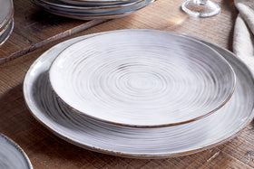 Two plates from the nkuku Dhanuk Ceramic Dinnerware Set (Set of 12), featuring a swirling white glaze and brown edges, are stacked on a rustic wood table with a linen napkin and wine glass in the background.