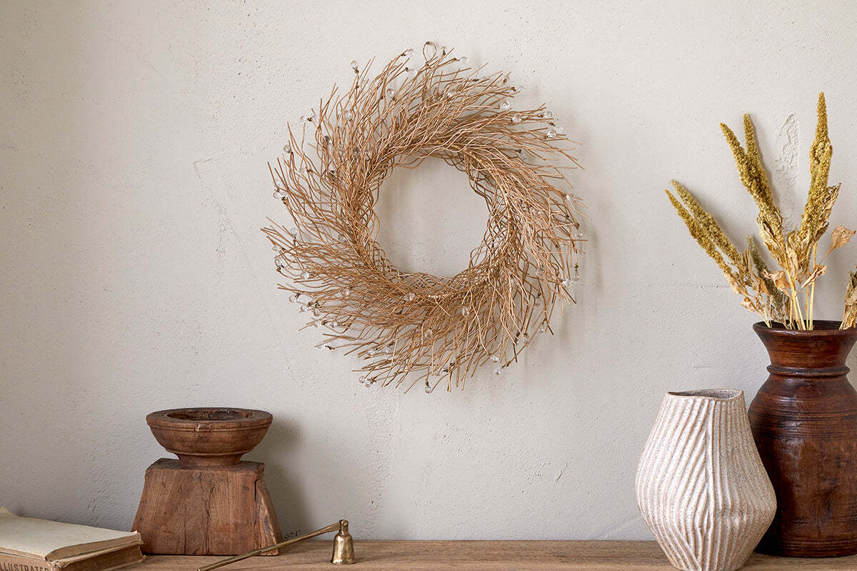 A minimalist scene showcases nkuku’s Danvi Glass Drop Wreath – Brass & Clear, hanging on a neutral wall above a rustic wood surface with a wooden bowl, striped ceramic vase, brown vase with dried grasses, brass bell, and closed book.