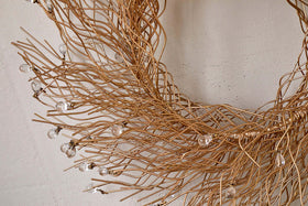 The Danvi Glass Drop Wreath by nkuku features intertwined brass wires shaped into a circle, with clear glass beads resembling dewdrops. Shown here against a plain, light beige wall.