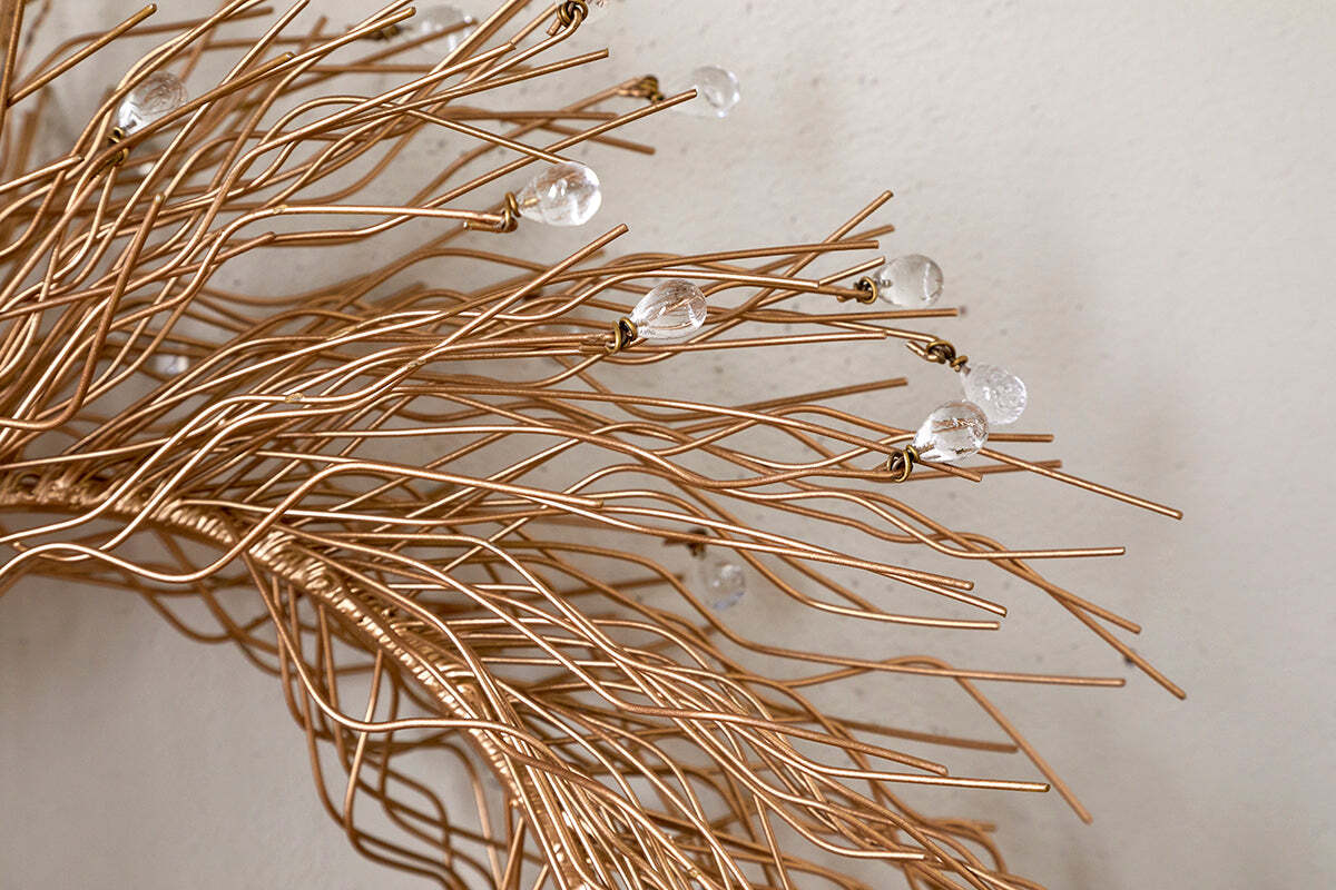 Close-up of the Danvi Glass Drop Wreath by nkuku, featuring intertwined brass wires with clear glass beads, creating a delicate, organic form against a light-colored wall.