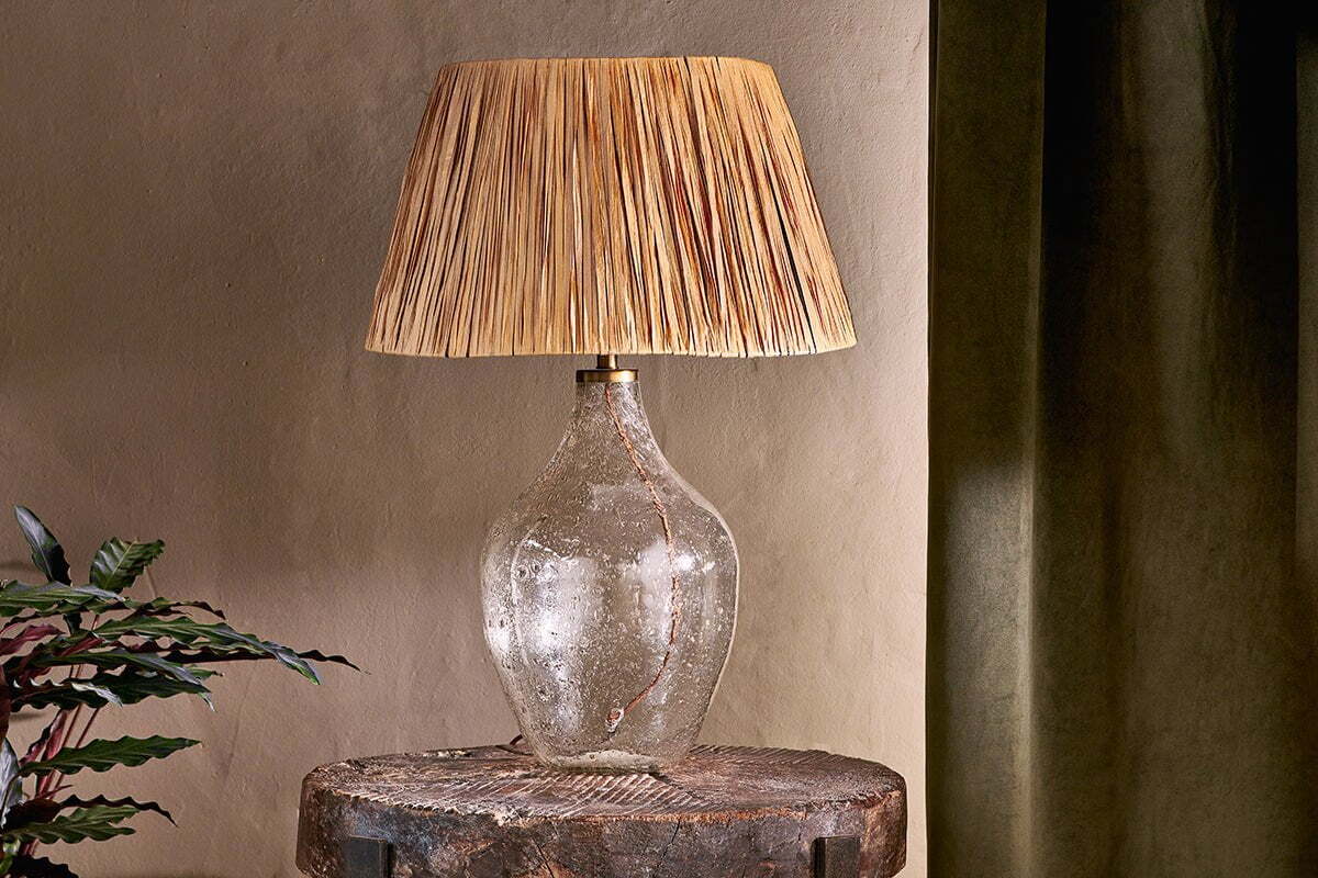 The nkuku Damini Recycled Glass Table Lamp, featuring a textured clear base and woven shade, is placed on a rustic wooden table beside a leafy plant, with a dark green curtain against a beige wall.