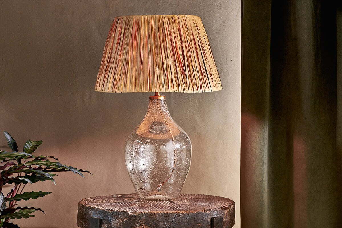 The Damini Recycled Glass Table Lamp by nkuku, featuring an organic recycled glass base and a straw-textured beige shade, sits on a rustic stump beside green leafy plants and dark curtains, set against a neutral wall.