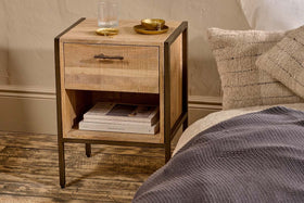 The Dasai Mango Wood Bedside Table by nkuku, featuring a drawer and open shelf, holds a glass of water, gold dish, and bowl. Books are stacked on the shelf beside a bed with a textured grey blanket and beige pillow on wooden flooring.