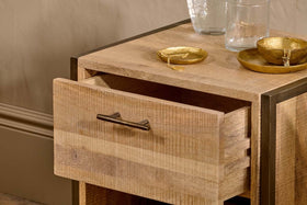 The Dasai Mango Wood Bedside Table by nkuku, featuring an open drawer with a simple metal handle, displays two gold bowls with jewelry and two clear glasses on top. Soft lighting highlights its handcrafted mango wood grain against a beige wall.