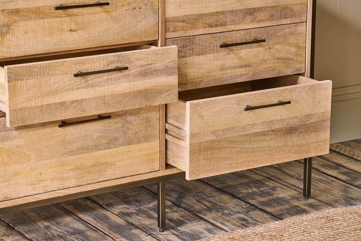 The Dasai Mango Wood Chest of Drawers by nkuku, featuring black metal handles and slender legs, stands on a wooden floor. Several drawers are ajar, displaying the rich grain. A beige wall and woven rug enhance the modern scene.