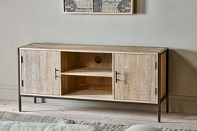 The nkuku Dasai Mango Wood Media Unit with black metal legs and handles stands against a light gray wall. It features two closed cabinets, two open shelves with cable cutouts, and has framed artwork displayed above.