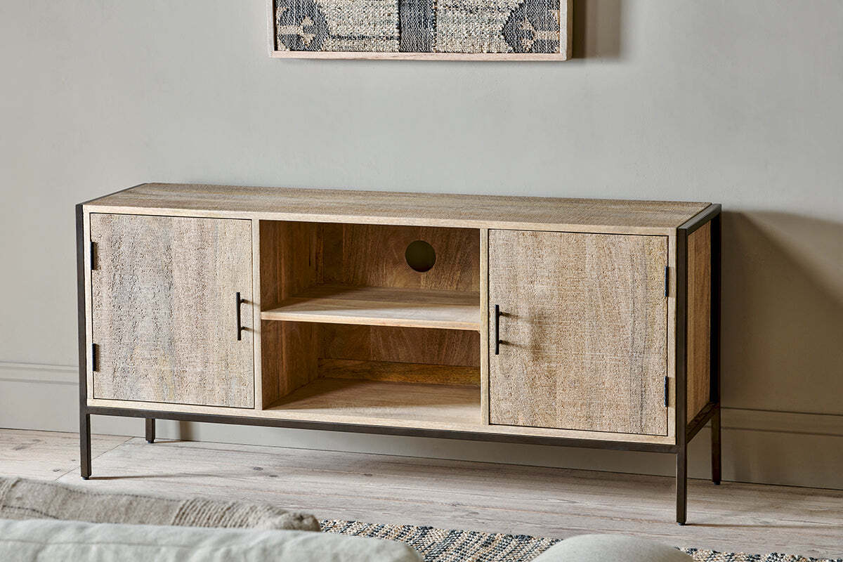 The nkuku Dasai Mango Wood Media Unit with black metal legs and handles stands against a light gray wall. It features two closed cabinets, two open shelves with cable cutouts, and has framed artwork displayed above.