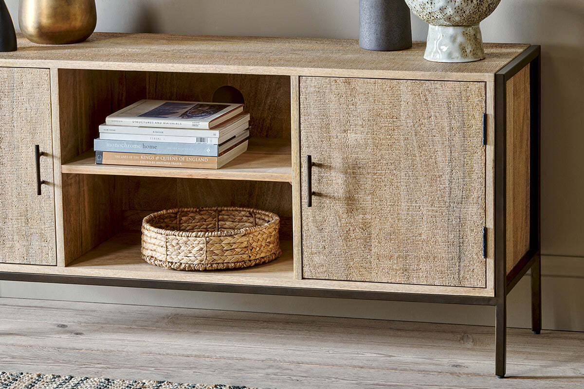 The Dasai Mango Wood Media Unit by nkuku serves as a stylish entertainment console, featuring two closed cabinets, an open shelf with books and a basket, black metal legs, and decorative items atop its light mango wood finish.
