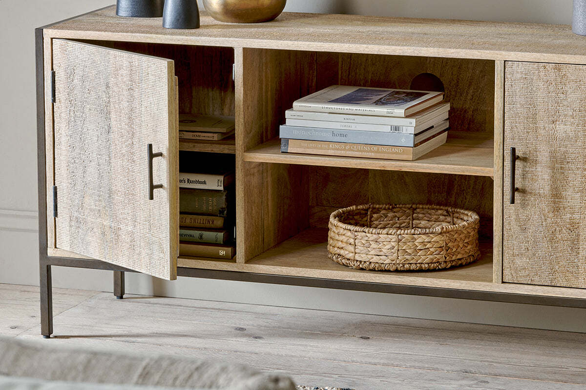 The nkuku Dasai Mango Wood Media Unit features light mango wood with open shelving displaying books and a woven basket on the lower shelf.