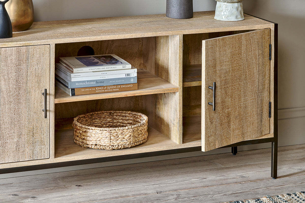 The Dasai Mango Wood Media Unit by nkuku features light mango wood with black handles, set on a wood floor. One door is open to show two shelves with stacked books and a woven basket. Decorative objects adorn this stylish console.