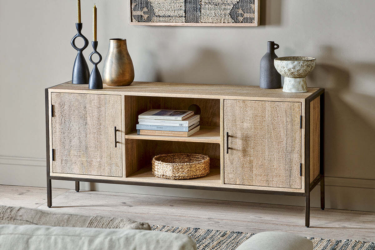 The Dasai Mango Wood Media Unit by nkuku, featuring black metal legs, stands against a neutral wall. Books and a woven basket rest on its center shelf, while decorative vases, candlesticks, and a ceramic bowl adorn the top. A textured rug and artwork peek into view.