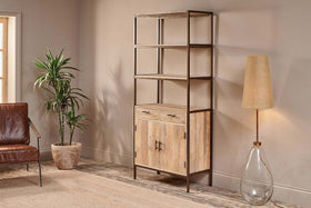 A modern living room showcases the nkuku Dasai Mango Wood Storage Unit, featuring drawers and cabinet doors for stylish storage. To the left sits a potted plant and brown leather chair; to the right is a glass-base floor lamp with beige shade.