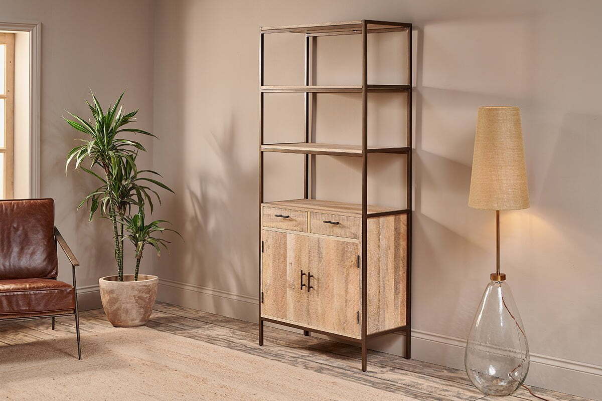 A modern living room showcases the nkuku Dasai Mango Wood Storage Unit, featuring drawers and cabinet doors for stylish storage. To the left sits a potted plant and brown leather chair; to the right is a glass-base floor lamp with beige shade.