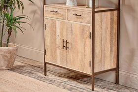 The Dasai Mango Wood Storage Unit by nkuku stands against a beige wall, offering stylish storage with two upper drawers, double doors, and dark metal frame accents. Sustainable and modern, it complements a potted plant and beige woven rug in the living room.