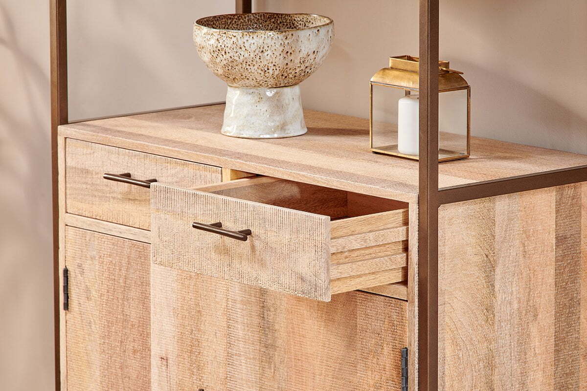 The nkuku Dasai Mango Wood Storage Unit features two doors and two drawers, one partially open. A speckled ceramic bowl and gold lantern with a white candle rest on top—stylish storage for your living room against a beige wall.