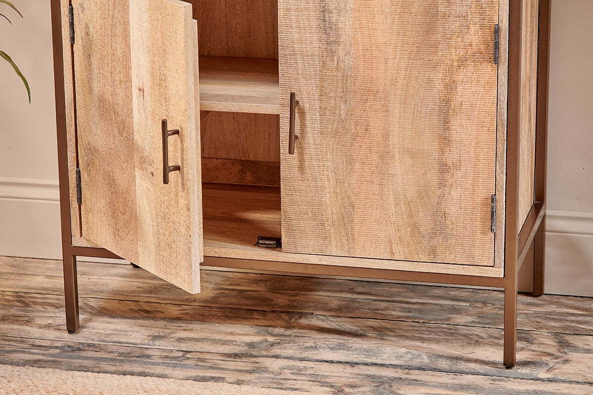 The Dasai Mango Wood Storage Unit by nkuku features two doors—one partially open showing an interior shelf—a natural mango wood grain finish, metal handles, slim metal legs, and stands on a textured wood-effect floor.