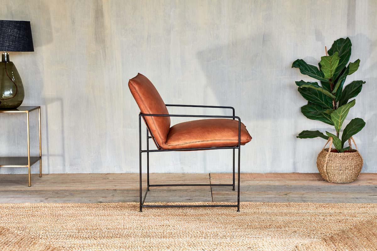 The Durium Leather Lounger - Aged Tan by nkuku, featuring a modern iron frame, rests on a woven rug. To the left sits a glass lamp on a gold table; to the right is a potted plant in a basket, set against a textured wall and wooden floor.
