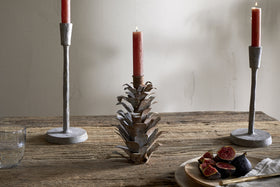 A rustic wooden table features two metal candlesticks, including the nkuku Elagalu Pine Cone Candle Holder in zinc. To the right is a wooden plate with sliced figs and cutlery, while a clear glass of water sits to the left against a plain wall.