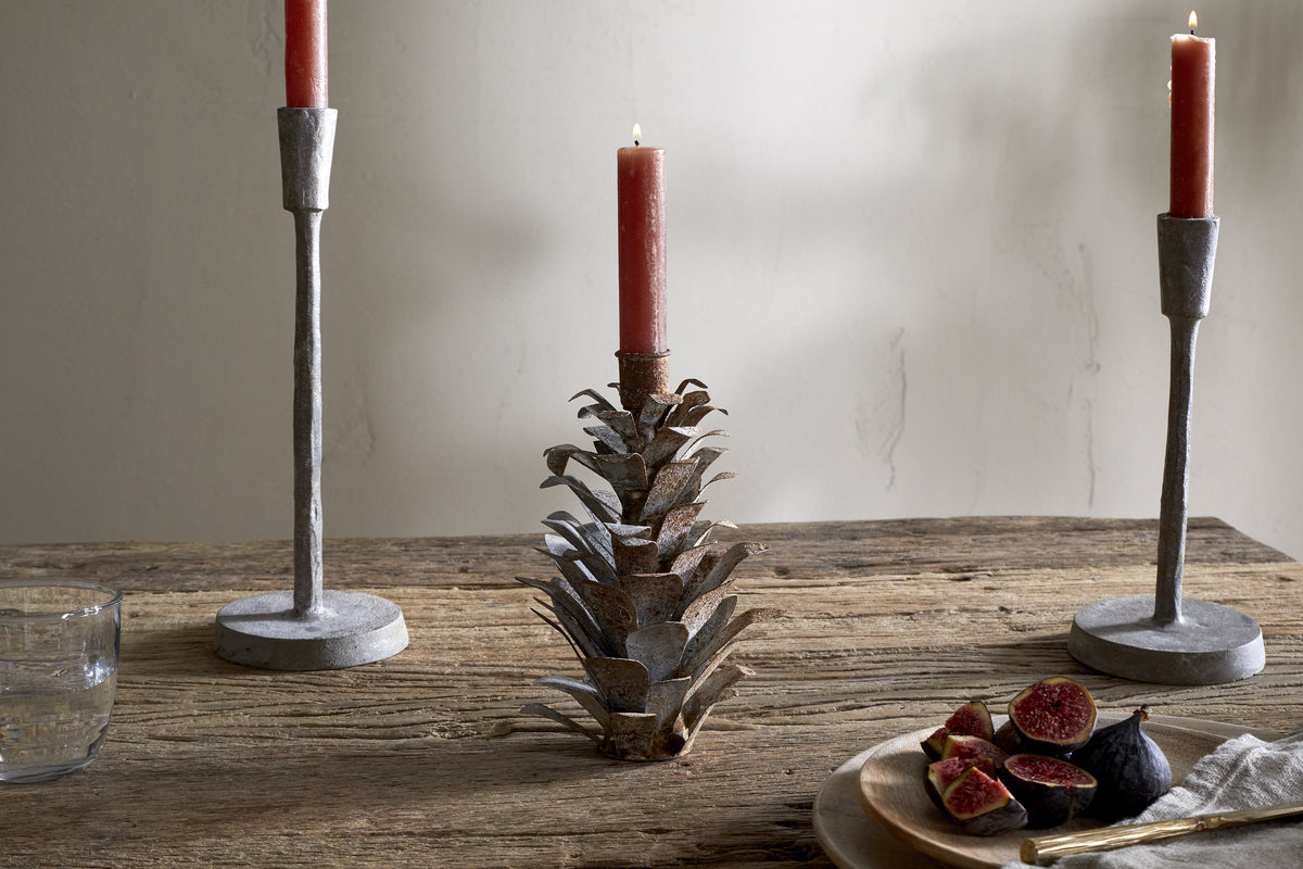 A rustic wooden table features two metal candlesticks, including the nkuku Elagalu Pine Cone Candle Holder in zinc. To the right is a wooden plate with sliced figs and cutlery, while a clear glass of water sits to the left against a plain wall.