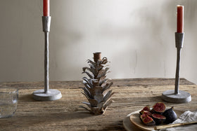 A rustic whitewashed wooden table displays two gray candlesticks with red candles, the nkuku Elagalu Pine Cone Candle Holder in zinc, a glass of water, and a wooden plate of figs against a neutral minimal background.