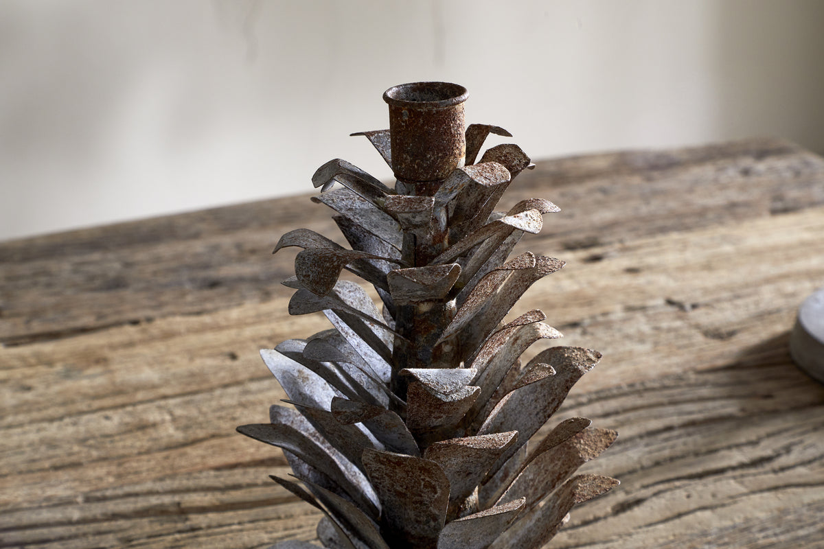 The nkuku Elagalu Pine Cone Candle Holder in zinc features a rustic, aged metal design, creating inviting festive decor. The pine cone shape sits beautifully on weathered wood, serving as a charming addition to any tabletop setting.