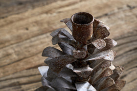 The nkuku Elagalu Pine Cone Candle Holder – Zinc features a textured, weathered metal design in brown and gray tones, displayed on a wooden surface with blurred festive decor and natural wood grain in the background.