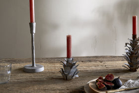 A rustic wooden table features nkuku's Elagalu Pine Cone Candle Holders - Zinc (Set of 2), with red candles, a glass of water, a beige cloth, and sliced figs on a wooden plate. The softly lit wall completes the festive scene.
