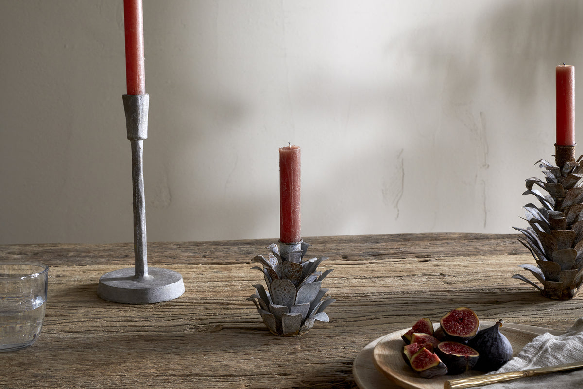 A rustic wooden table features nkuku's Elagalu Pine Cone Candle Holders - Zinc (Set of 2), with red candles, a glass of water, a beige cloth, and sliced figs on a wooden plate. The softly lit wall completes the festive scene.