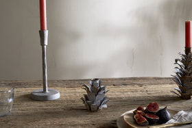 A rustic wooden table displays nkuku's Elagalu Pine Cone Candle Holders - Zinc (Set of 2), paired with a red candle, glass tumbler, metal artichoke accent, and a plate of halved figs and knife—all softly illuminated for festive charm.