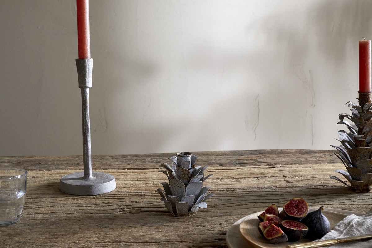 A rustic wooden table displays nkuku's Elagalu Pine Cone Candle Holders - Zinc (Set of 2), paired with a red candle, glass tumbler, metal artichoke accent, and a plate of halved figs and knife—all softly illuminated for festive charm.