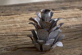 The nkuku Elagalu Pine Cone Candle Holders - Zinc (Set of 2) feature layered, metallic petals and a rustic look, making them ideal as festive tabletop decor or unique Christmas candleholders on a weathered wood surface.