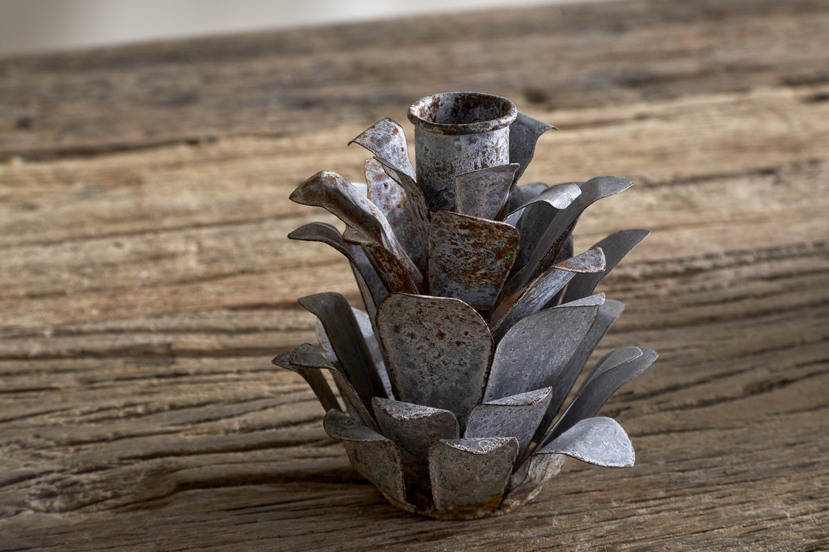 The nkuku Elagalu Pine Cone Candle Holders - Zinc (Set of 2) feature layered, metallic petals and a rustic look, making them ideal as festive tabletop decor or unique Christmas candleholders on a weathered wood surface.