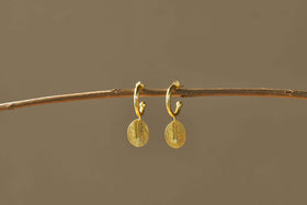The Esta Hoop Earrings - Gold by nkuku feature oval, hammered pendants hanging from small hoops. Shown on a thin brown branch against a beige background, their minimal design adds an elegant touch. One Size.