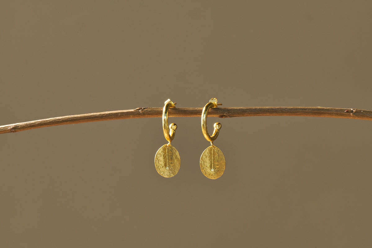 The Esta Hoop Earrings - Gold by nkuku feature oval, hammered pendants hanging from small hoops. Shown on a thin brown branch against a beige background, their minimal design adds an elegant touch. One Size.