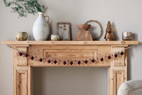 A wooden mantel displays vintage style with nkuku’s Ekiti Bauble Garland - Ruby & Copper, rustic candles, a white textured vase with greenery, framed art, a wooden bowl, beaded wreath, and metallic tree figurine against a light beige wall.