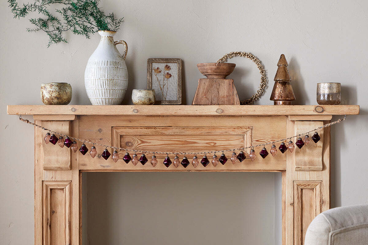 A wooden mantel displays vintage style with nkuku’s Ekiti Bauble Garland - Ruby & Copper, rustic candles, a white textured vase with greenery, framed art, a wooden bowl, beaded wreath, and metallic tree figurine against a light beige wall.