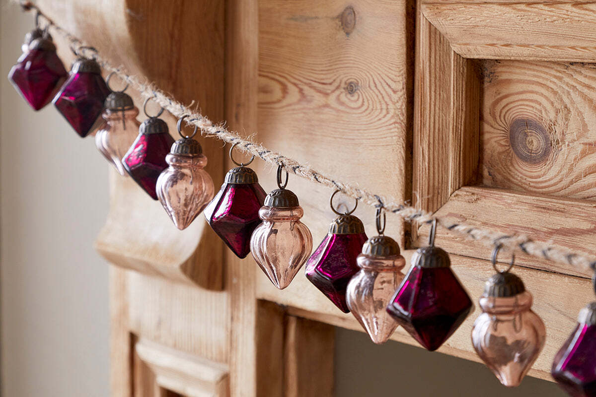 The Ekiti Bauble Garland - Ruby & Copper by nkuku features geometric, recycled glass ornaments with metal caps, strung on twine for a festive, vintage look—perfect for adding cozy charm to your mantelpiece.