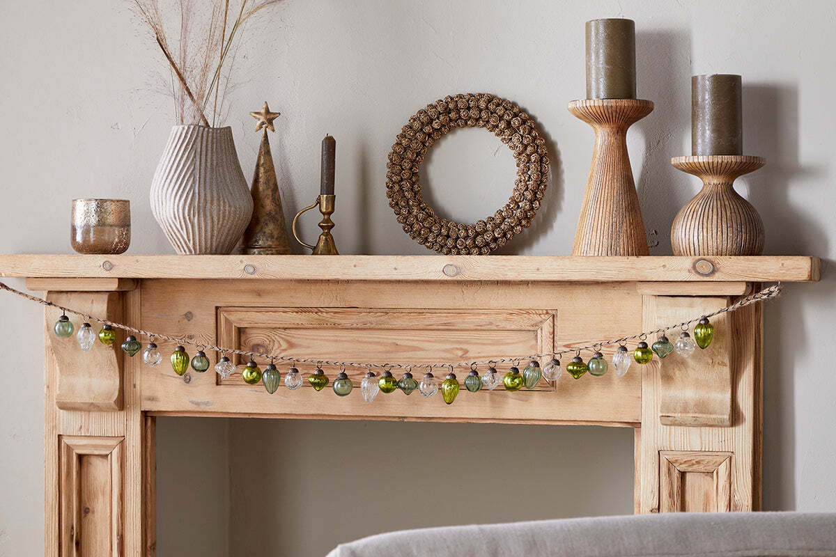 A light wooden mantelpiece decorated with vases, candles, a round beaded mirror, and the nkuku Ekiti Bauble Garland in mixed green and clear. Textured ceramics, recycled glass, and dried grasses add cozy festive charm.
